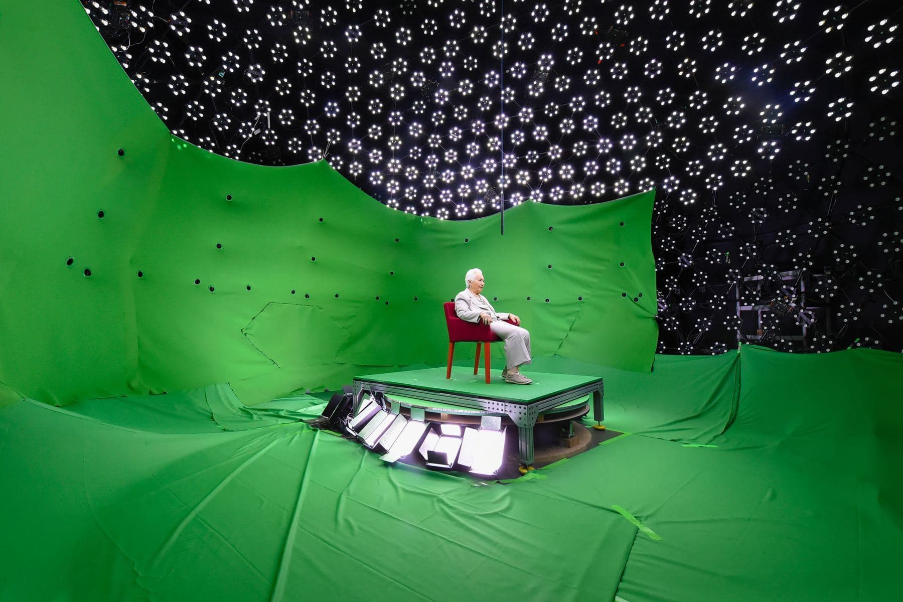 Renée Firestone seated in a red chair on a green-screen stage beneath a dome of lights during the Dimensions in Testimony recording