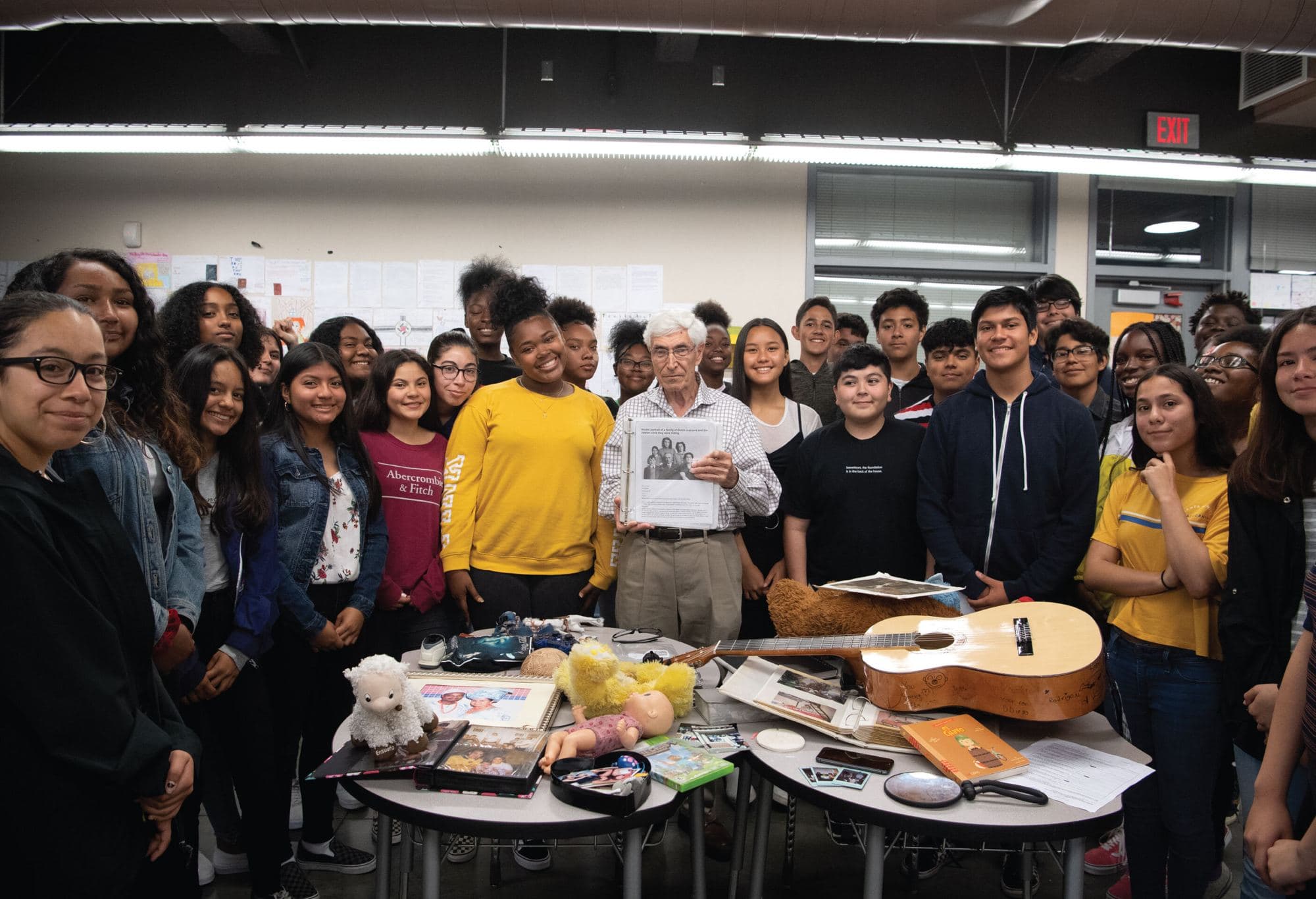 A classroom of students gathered around a Holocaust survivor holding a family photograph, with artifacts and a guitar on the tables in front of them