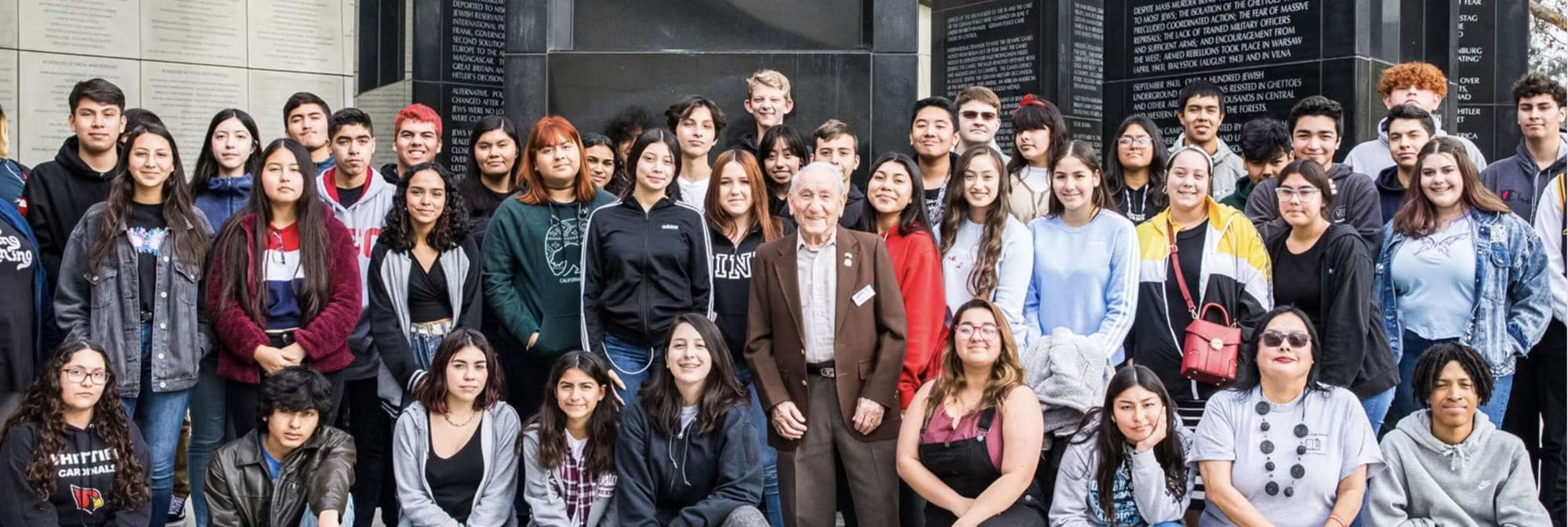 A Holocaust survivor stands with a visiting high school class in front of the memorial wall